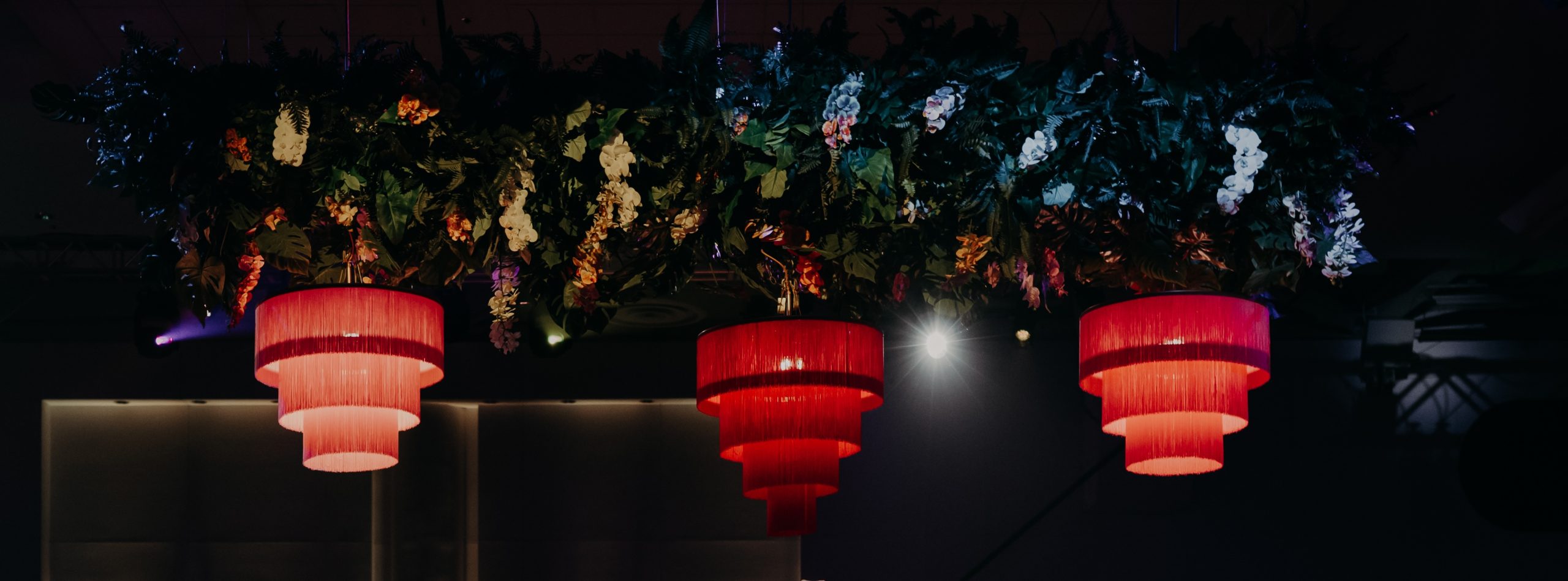 Elegant overhead décor featuring lush artificial greenery, cascading tropical flowers, and bold red chandeliers at a themed event.