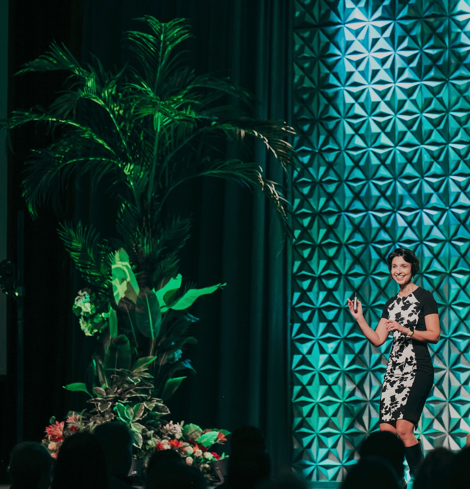 A speaker presenting on stage beside a dramatic tropical vignette with lush faux greenery and floral accents.