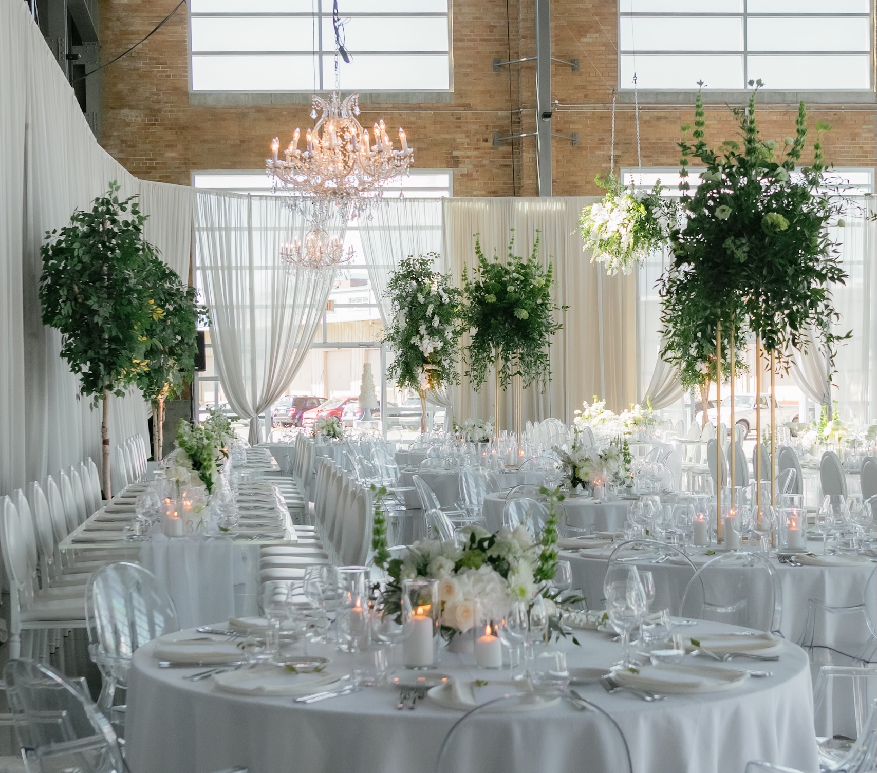 A luxurious wedding reception setup featuring artificial trees, elevated greenery centerpieces, and crystal chandeliers, creating an elegant and romantic atmosphere.