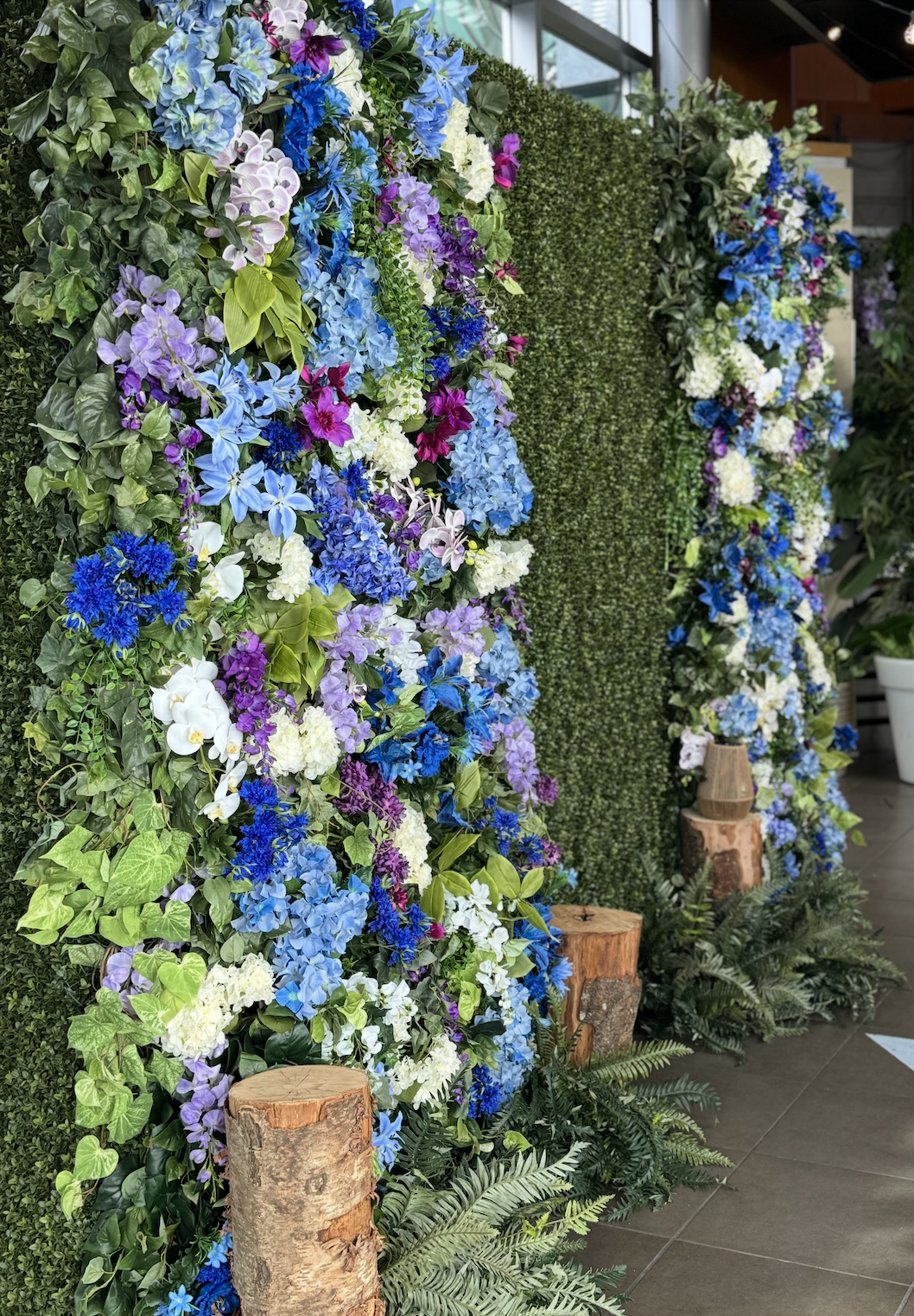 Lush faux floral wall backdrop with cascading blue, purple, and white flowers against green boxwood, accented with wooden logs and ferns for event rentals.