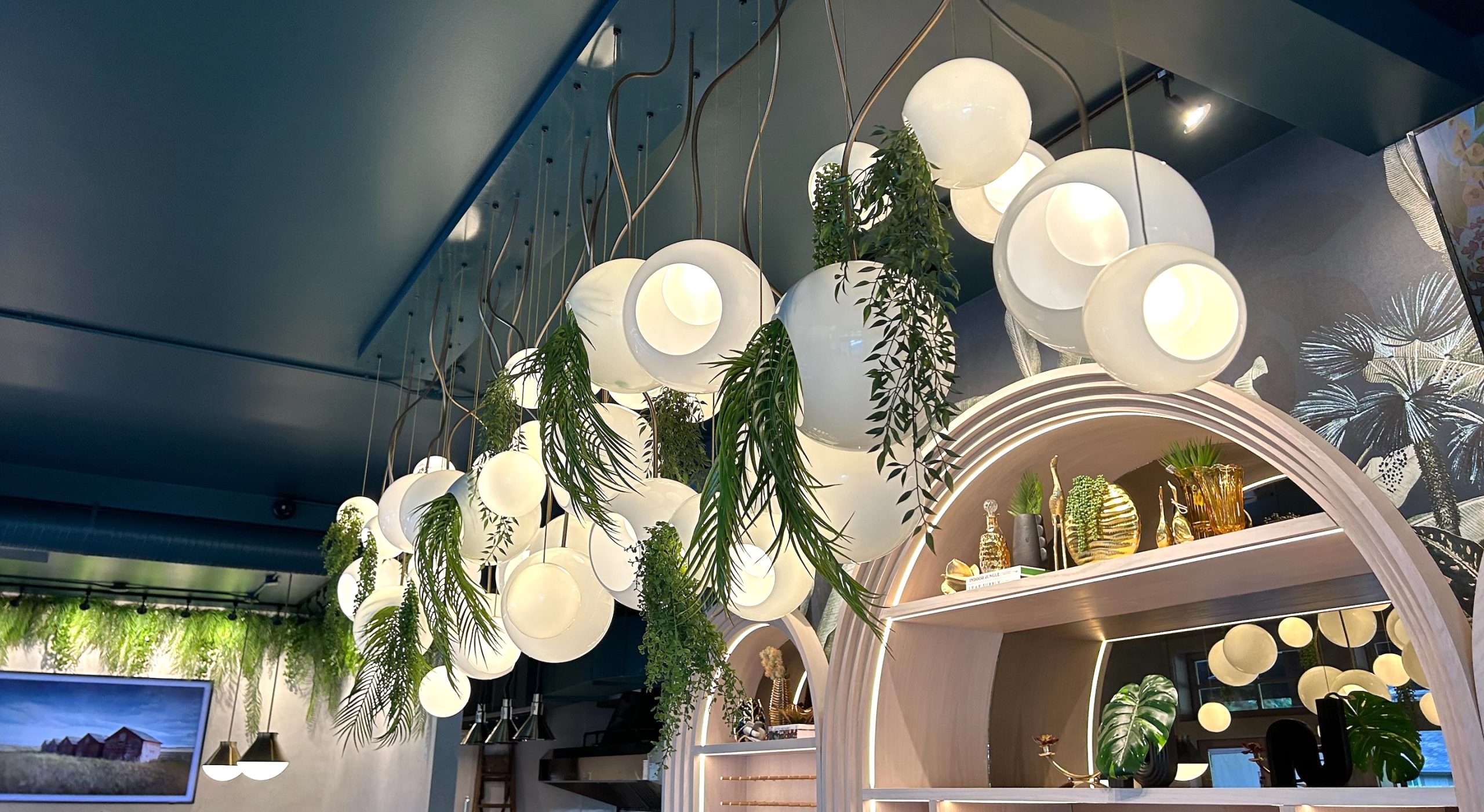 A modern restaurant chandelier adorned with cascading artificial greenery, featuring spherical white pendant lights and lush trailing foliage.