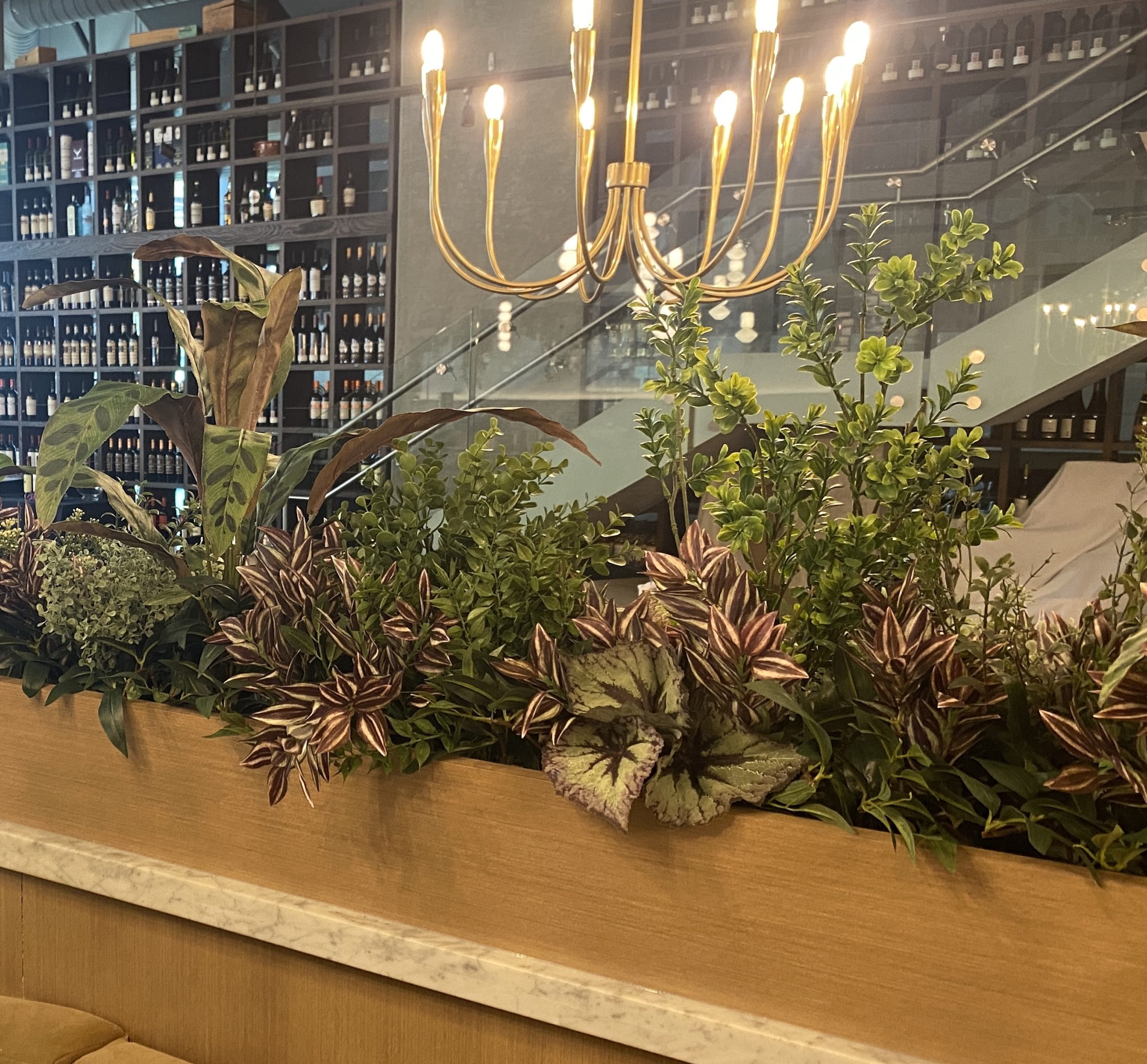 Artificial mixed greenery privacy dividers in a restaurant, featuring lush foliage in a wooden planter beneath a chandelier.