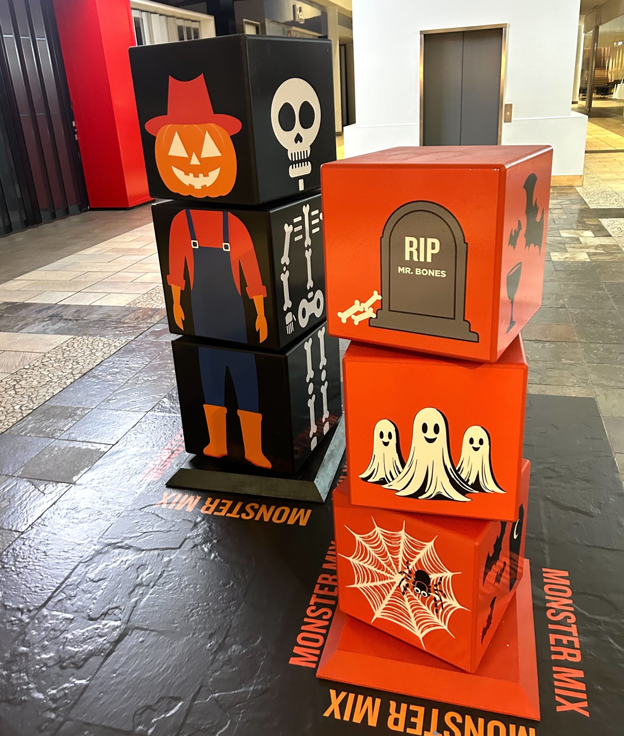 Stacked Halloween-themed cubes with playful illustrations of a pumpkin-headed scarecrow, skeleton, ghosts, and tombstones at The City of Lougheed Shopping Centre.