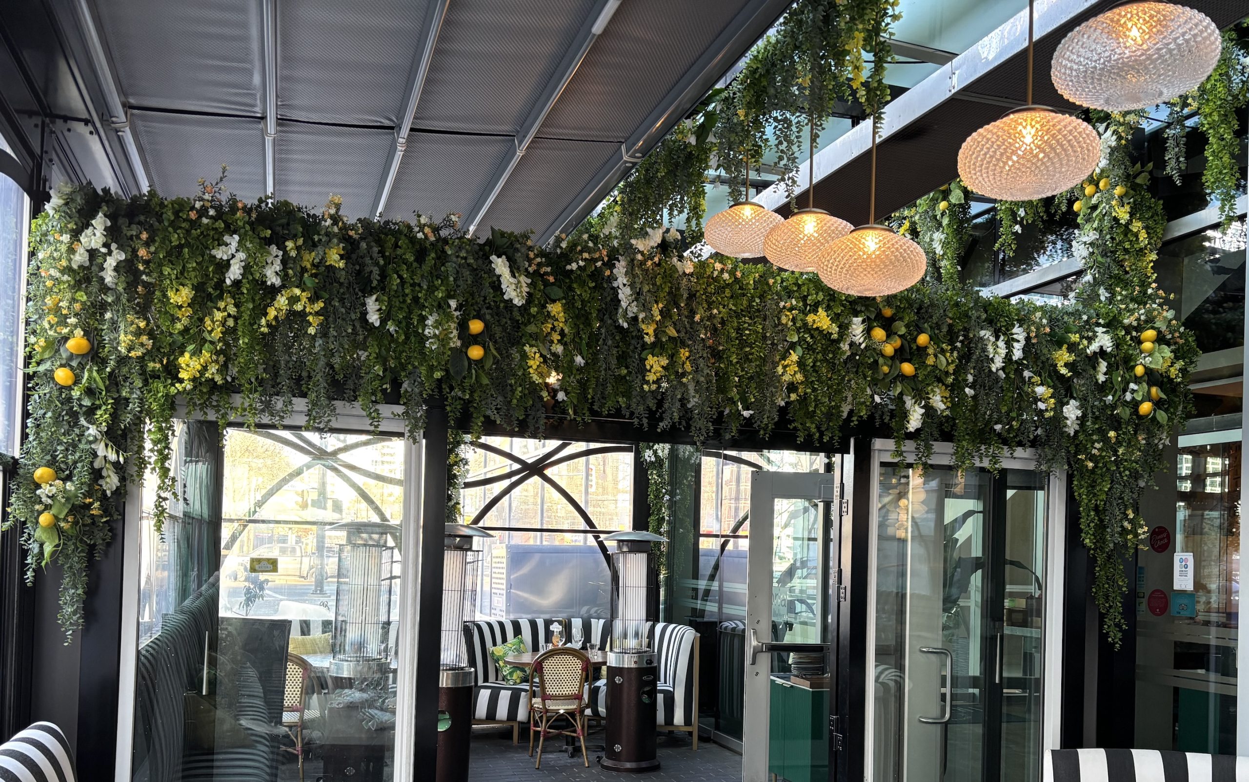 A vibrant exterior artificial overhead greenery installation featuring cascading greenery, white and yellow florals, and artificial lemons, creating a lush and inviting restaurant entrance.