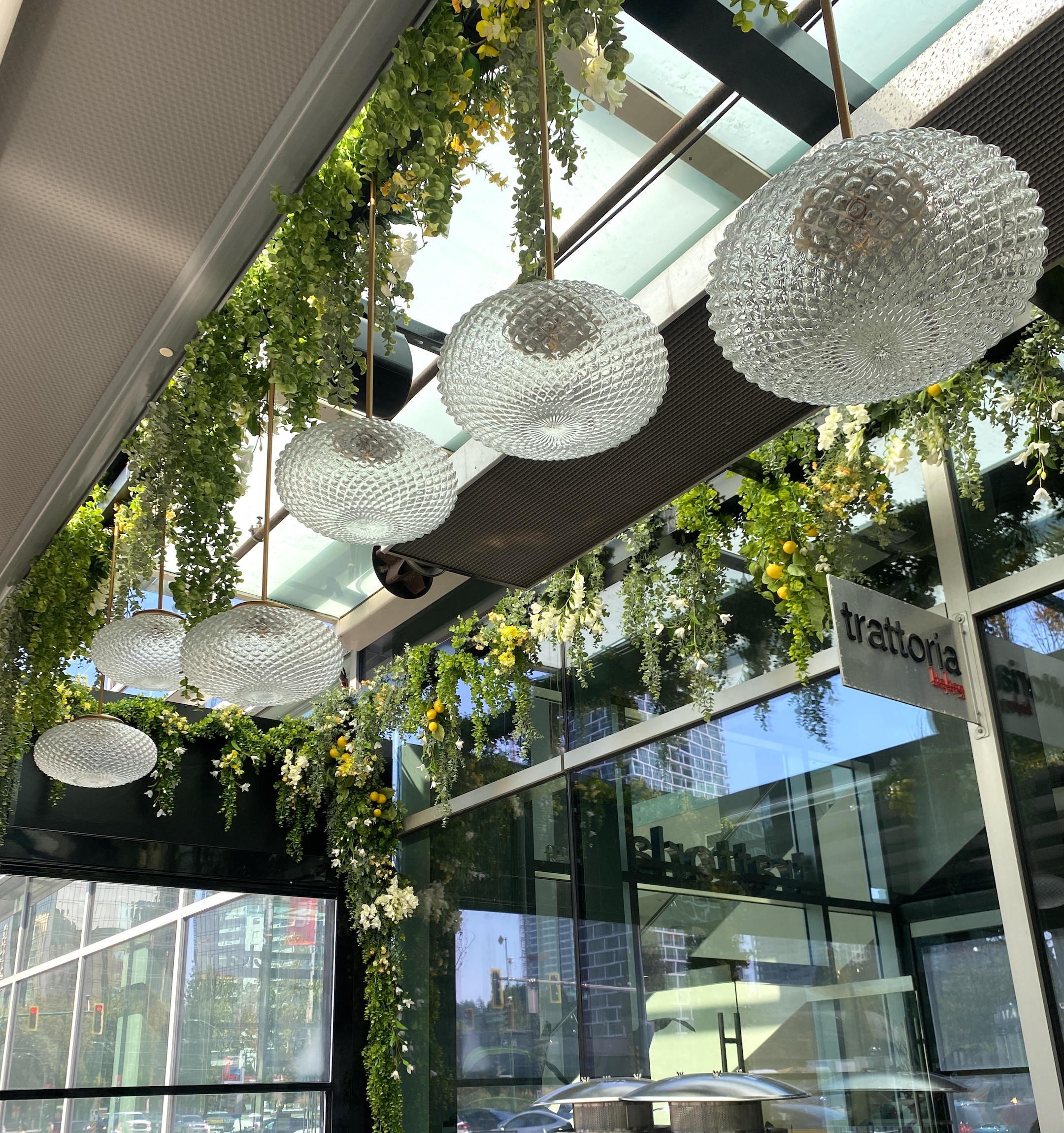 Faux trailing overhead greenery with cascading vines and lemon accents, enhancing the ambiance of a glass-roofed restaurant with elegant pendant lighting at Trattoria Restaurant in Burnaby.