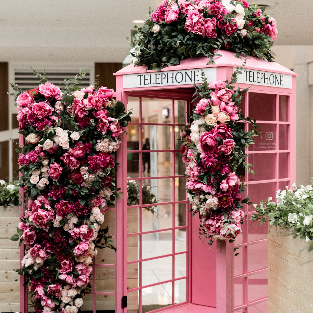 A vibrant pink telephone booth adorned with lush artificial flowers in shades of pink and blush, creating a stunning floral photo op installation.