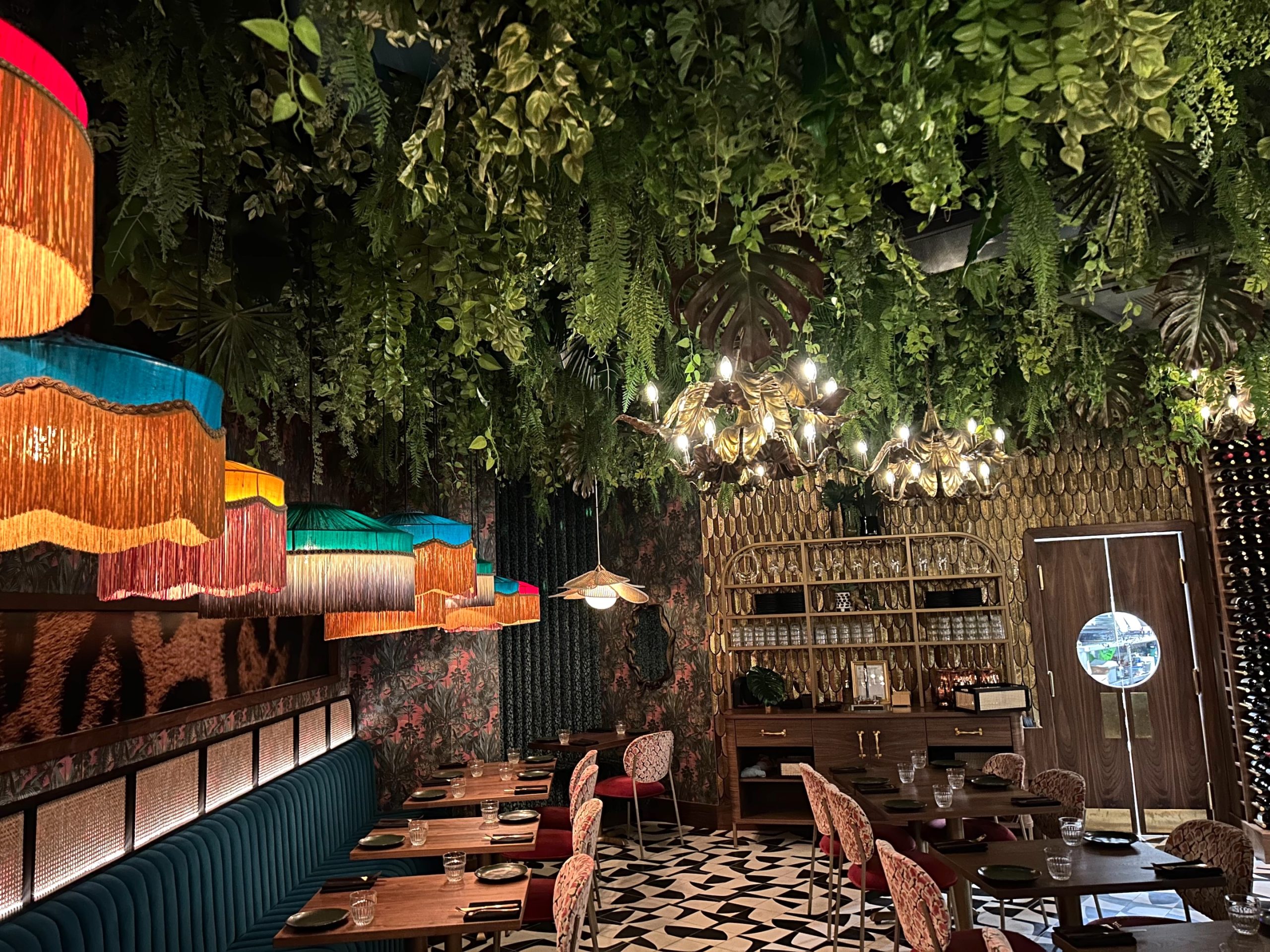 A restaurant with artificial ceiling greenery featuring cascading vines, ferns, and tropical leaves, complemented by colourful vintage-style pendant lighting.