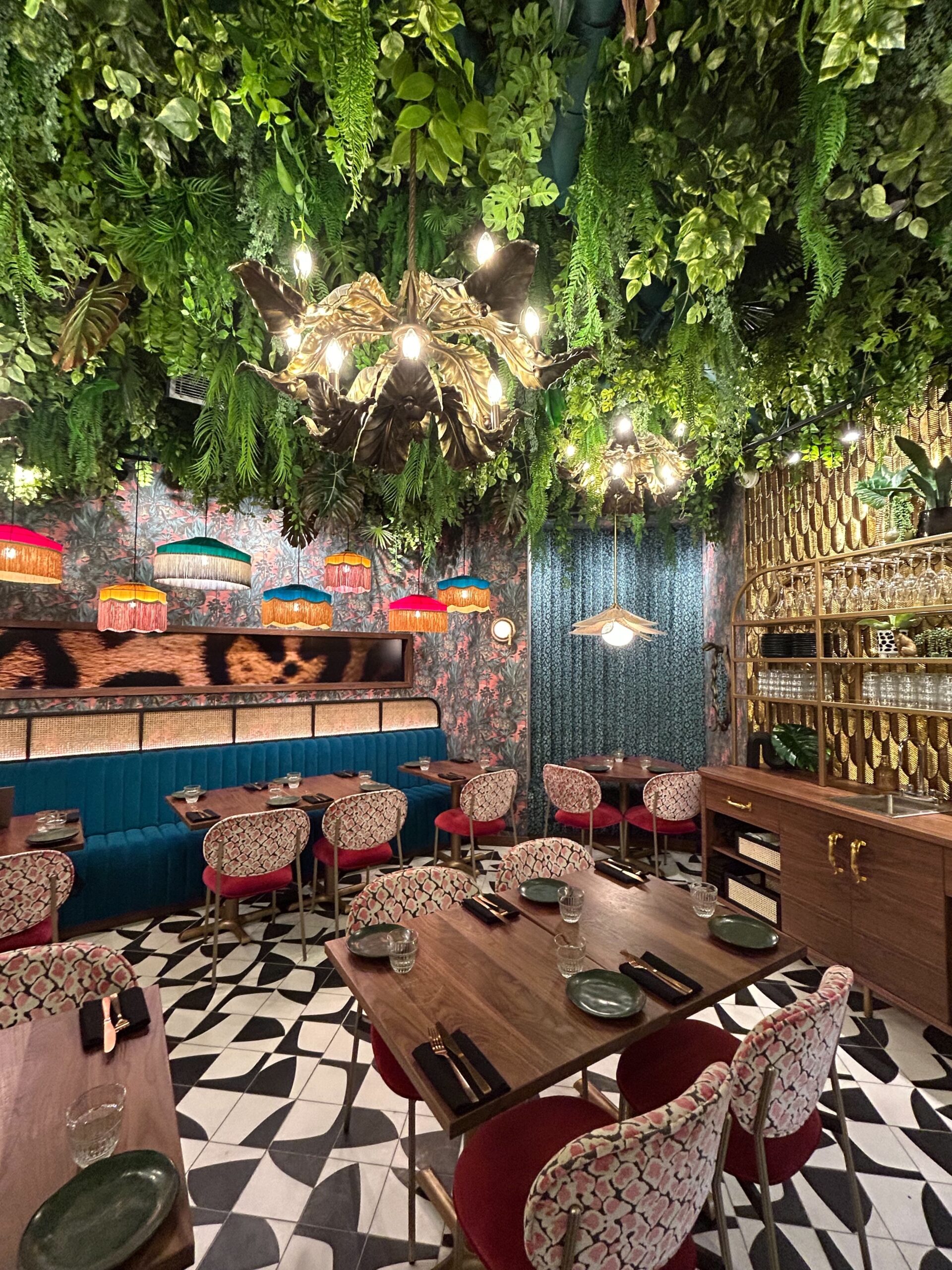 A restaurant interior featuring artificial ceiling greenery with cascading vines, ferns, and tropical leaves, enhancing a bold, jungle-inspired aesthetic.