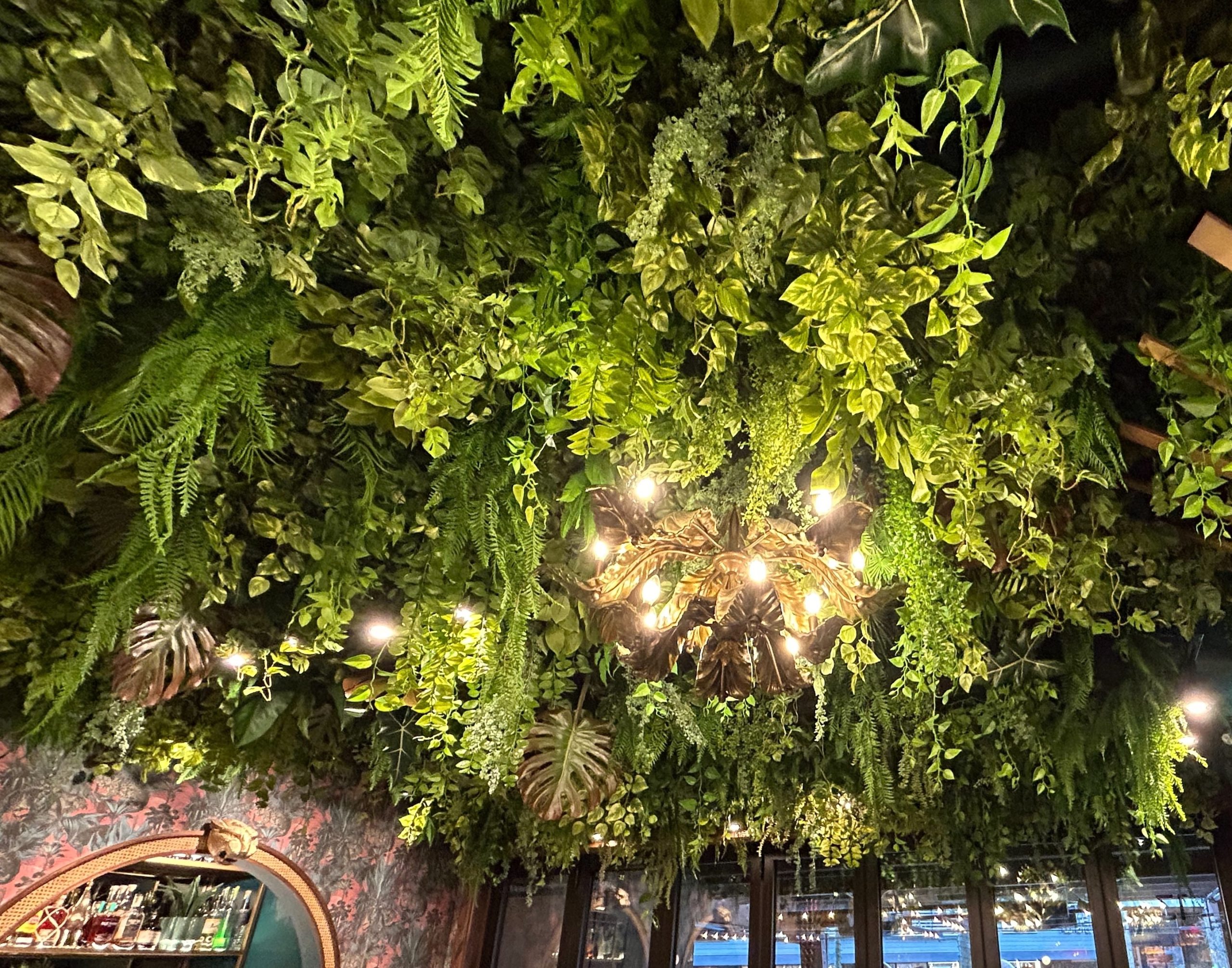 A restaurant ceiling covered in artificial greenery ceiling installation, including cascading vines, ferns, and tropical leaves, creating a lush, Latin America-inspired atmosphere