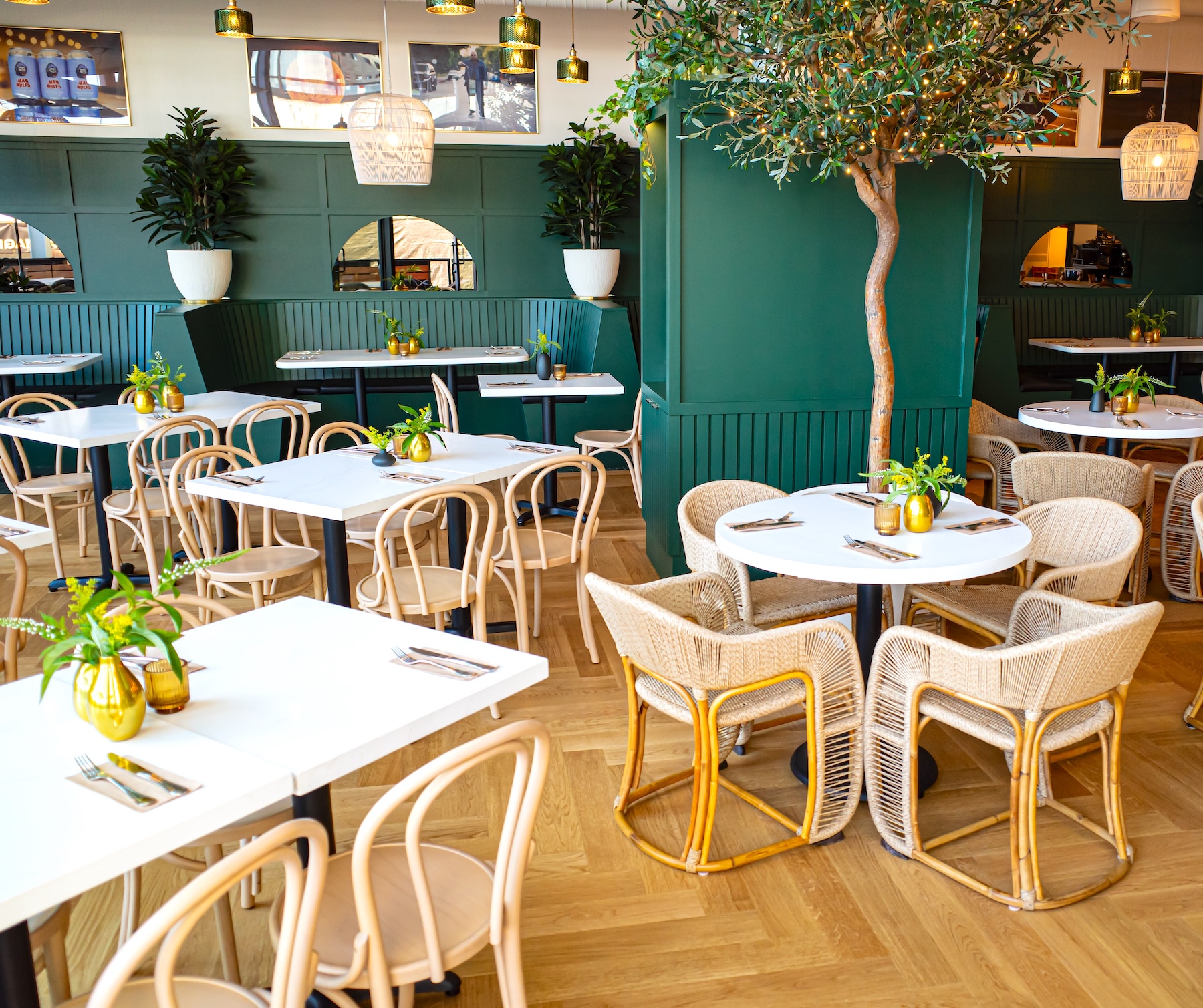 Artificial olive tree with warm string lights and faux potted plants enhance a stylish restaurant interior with green paneling, natural wood, and rattan seating.