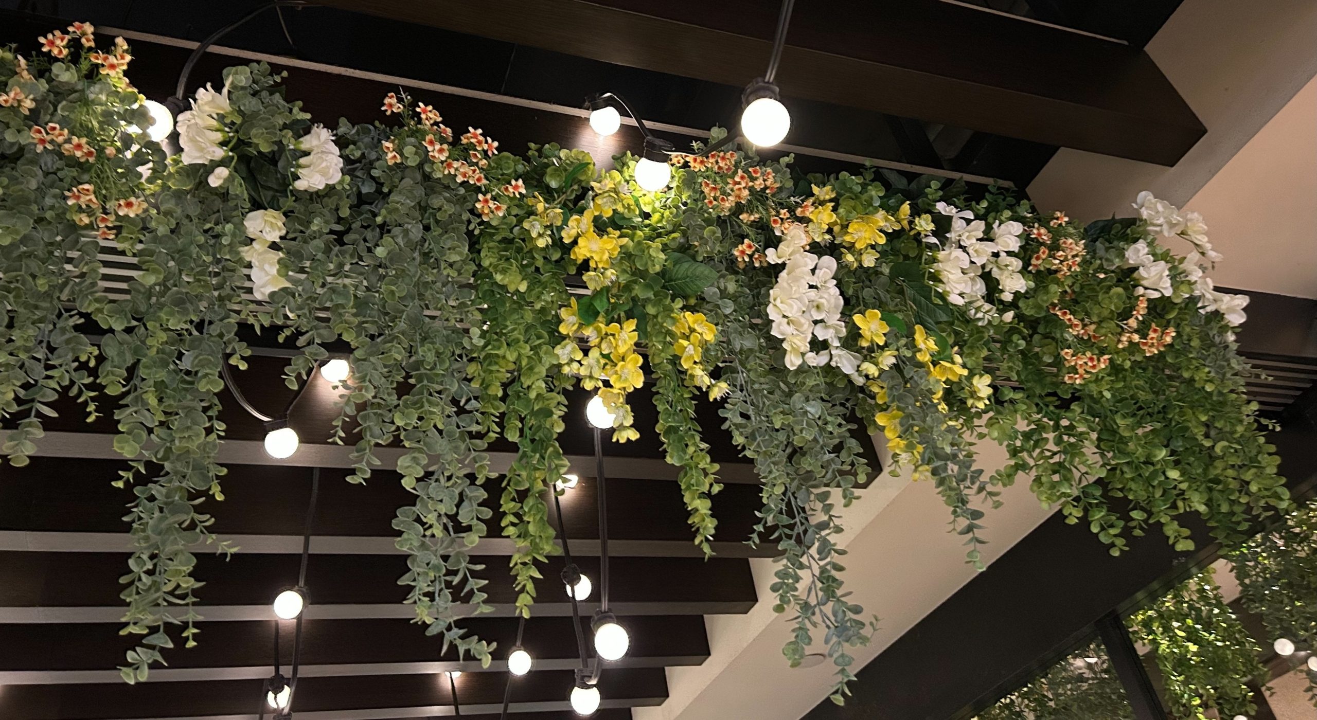 Lush artificial greenery with trailing vines, white orchids, and yellow flowers hanging from a restaurant ceiling, illuminated by warm overhead string lights.