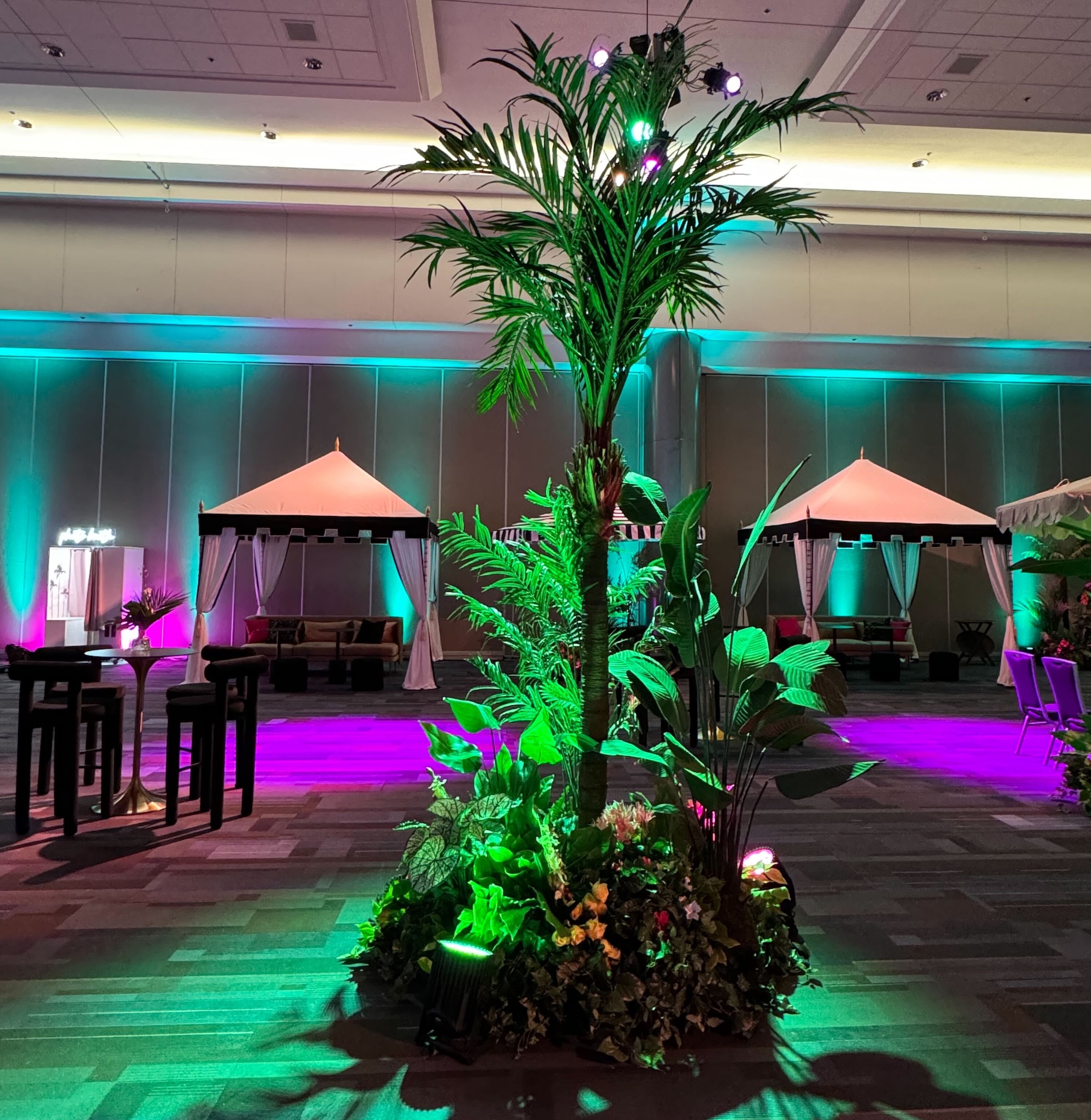 Artificial tropical palm tree and lush greenery centerpiece at a vibrant event, surrounded by cabanas and dramatic lighting in a modern venue.