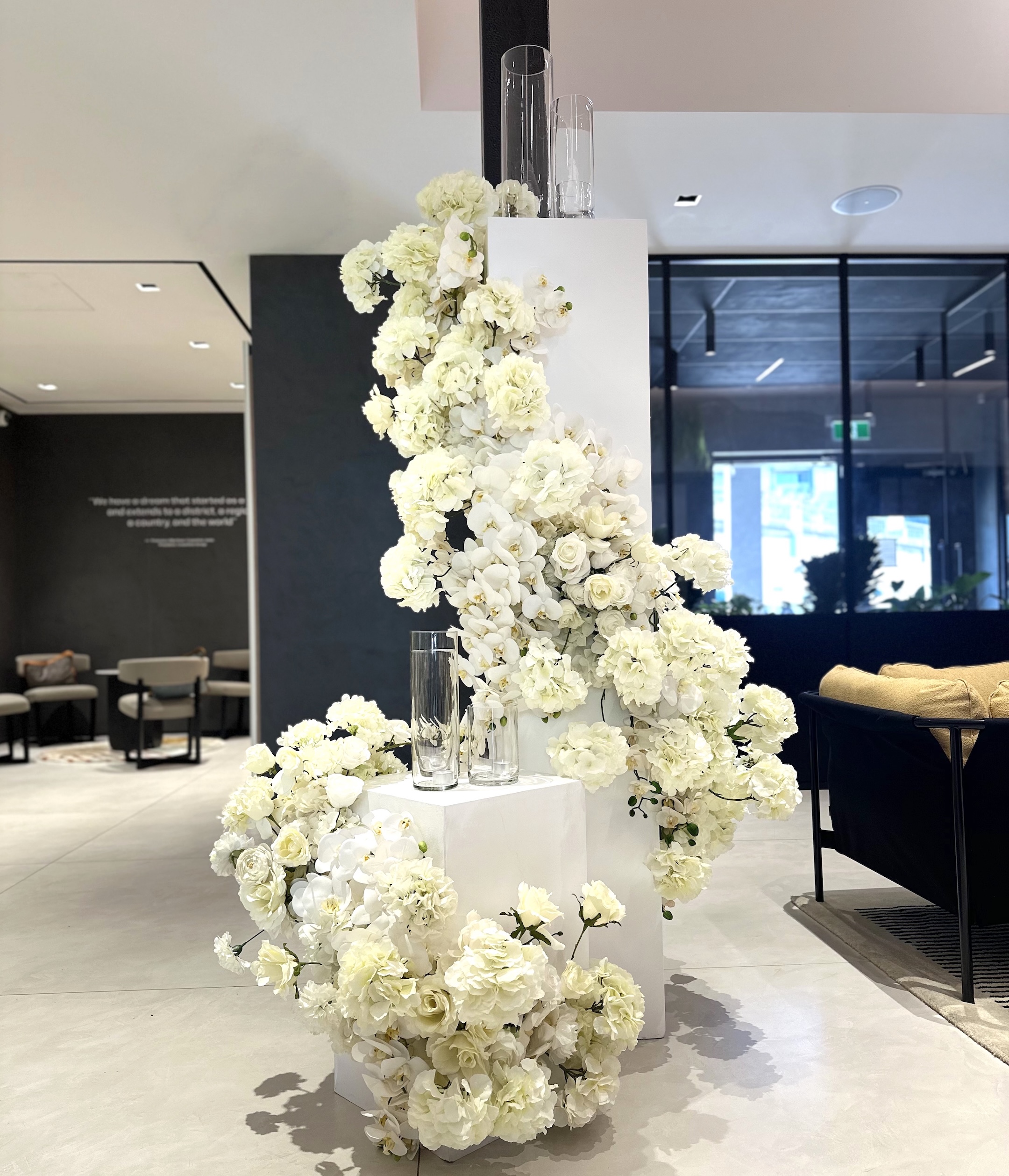 Luxurious cascading faux white floral arrangement with roses, orchids, and hydrangeas on tiered white pillars for an elegant event display.