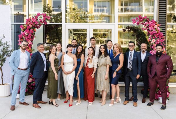 Group of people dressed in formal attire posing in front of a bright pink floral arch installation outside a building.