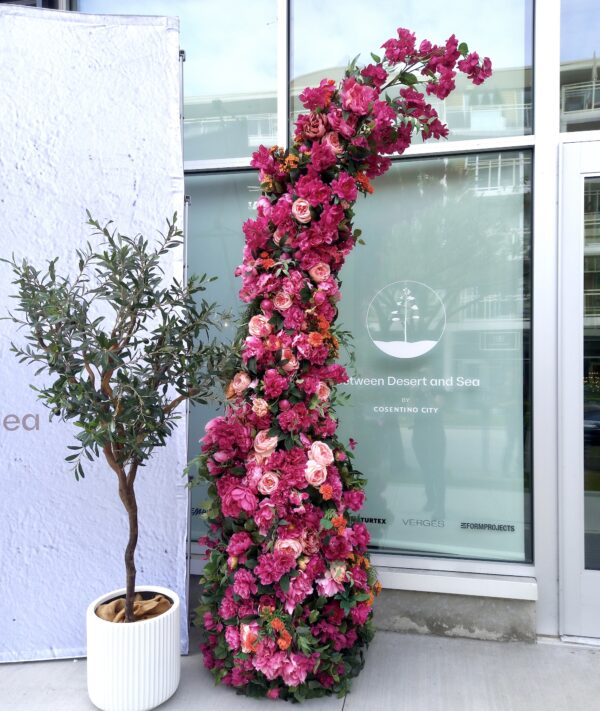 Close-up of one side of a vibrant pink and orange floral arch paired with a potted olive tree at an event entrance.
