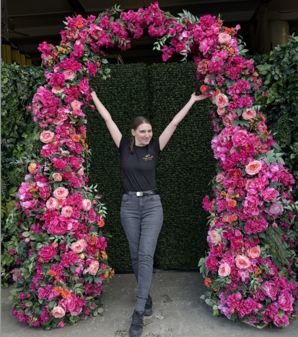 Person standing under a bold pink floral arch with arms raised, showcasing the installation against a green backdrop wall.