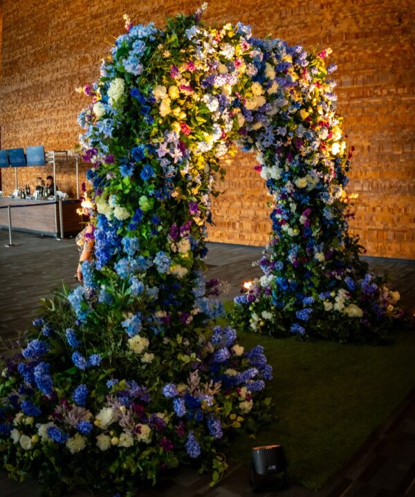 Lush floral entrance arch bursting with artificial blue, purple, and ivory blooms for a corporate event rental.