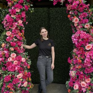 Team member standing beneath a vibrant floral arch with pink, coral, and orange artificial flowers in front of a green boxwood backdrop, created by Greenscape Design and Decor Inc.