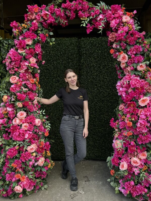 Team member standing beneath a vibrant floral arch with pink, coral, and orange artificial flowers in front of a green boxwood backdrop, created by Greenscape Design and Decor Inc.