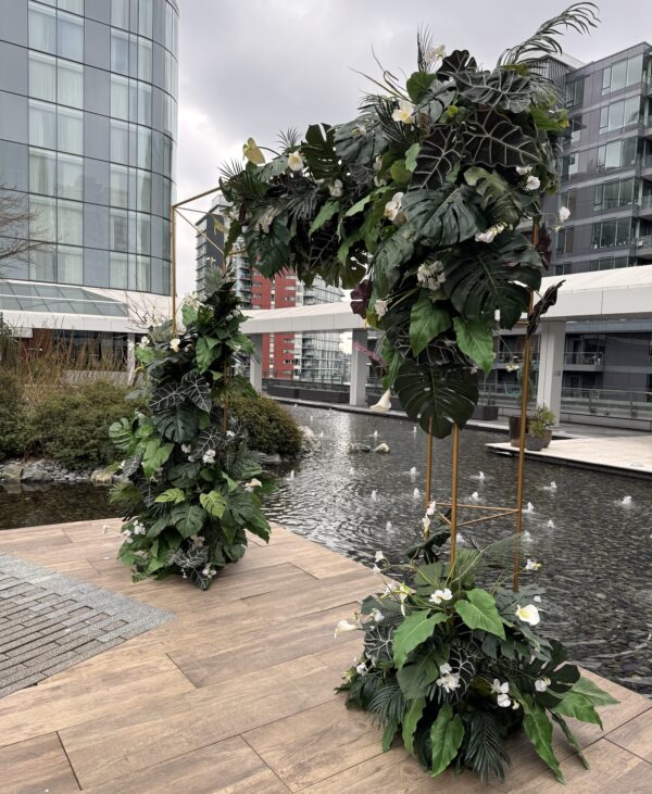 Modern tropical floral arch installation with greenery and orchids beside reflecting pool.