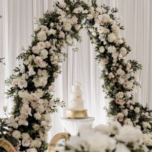 White floral wedding arch with lush greenery and soft blush, cream, and ivory blooms framing a tiered wedding cake on a gold stand.