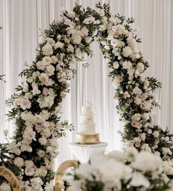 White floral wedding arch with lush greenery and soft blush, cream, and ivory blooms framing a tiered wedding cake on a gold stand.