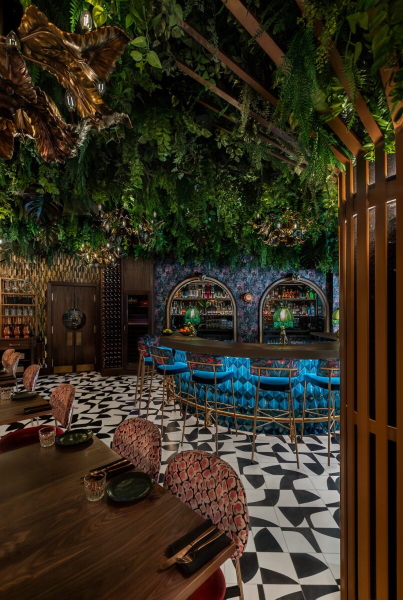 Restaurant bar with lush overhead greenery, patterned flooring, and bold jewel-toned seating for a dramatic look
