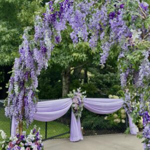 Purple faux wisteria climbing arch with cascading florals and draped fabric at an outdoor garden wedding ceremony.