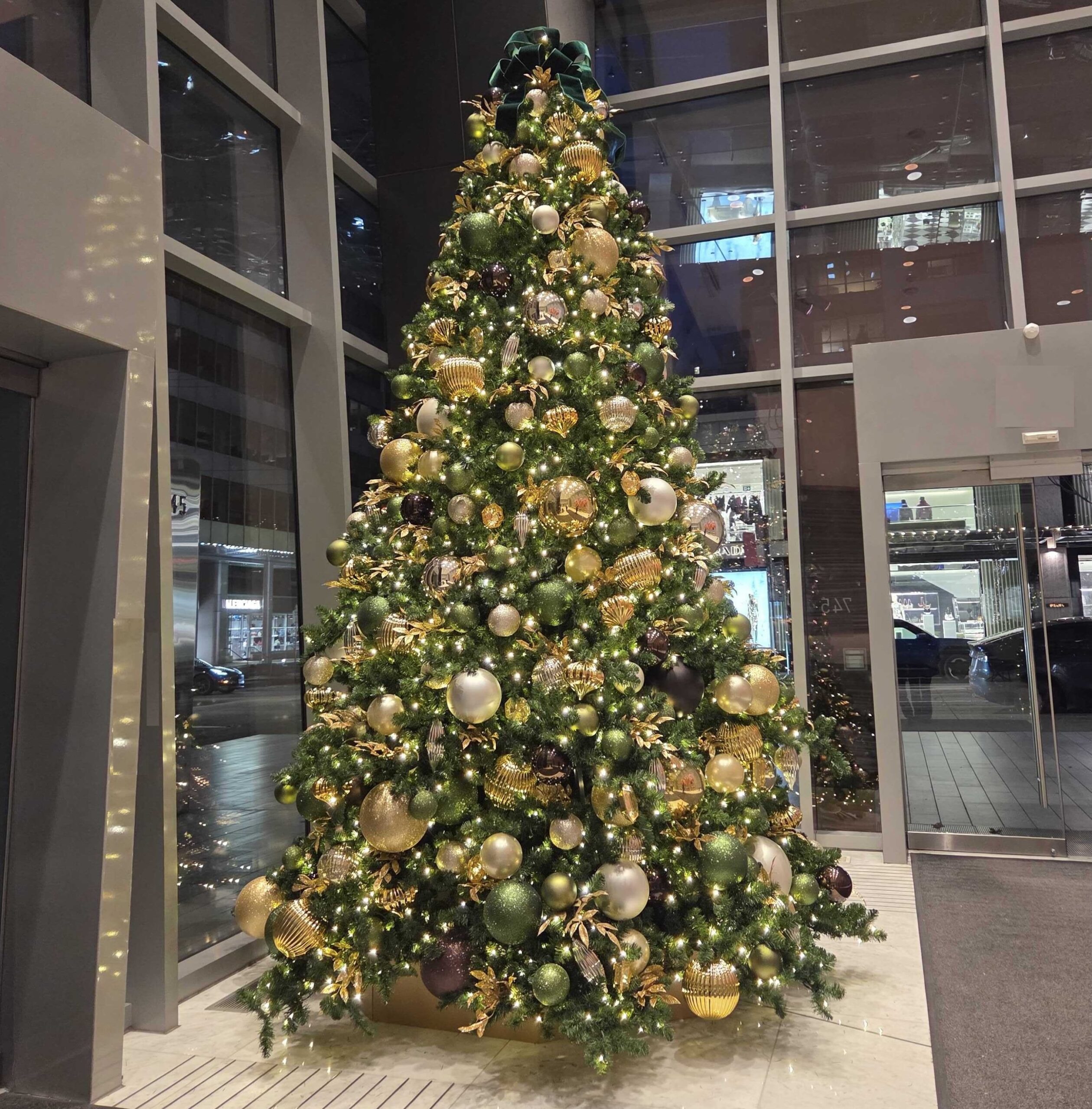 Large faux commercial Christmas tree with gold and green ornaments and warm white lights in a modern office lobby.