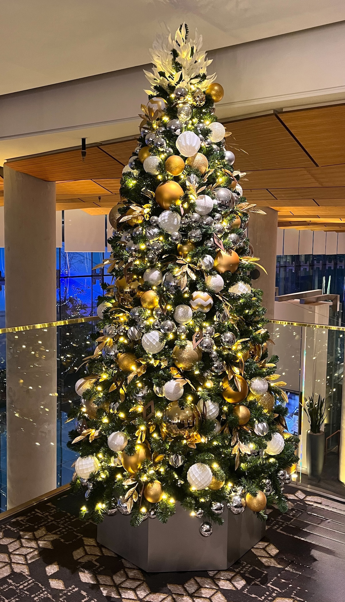 Large artificial Christmas tree decorated in gold, silver, and white with warm lighting, installed in a modern hotel lobby with glass railings.