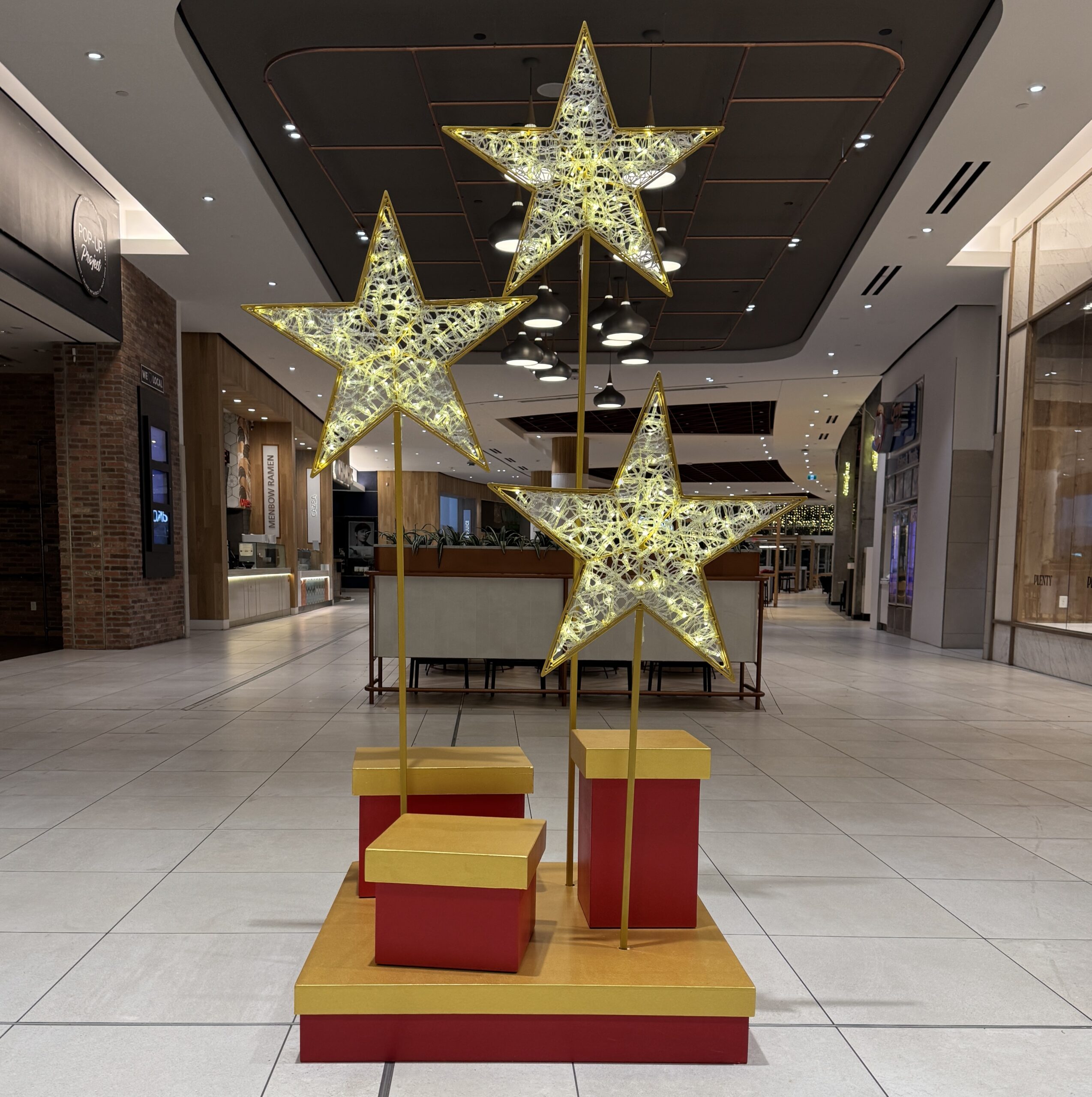 Illuminated gold star decorations mounted above red and gold gift boxes in a shopping centre holiday display.