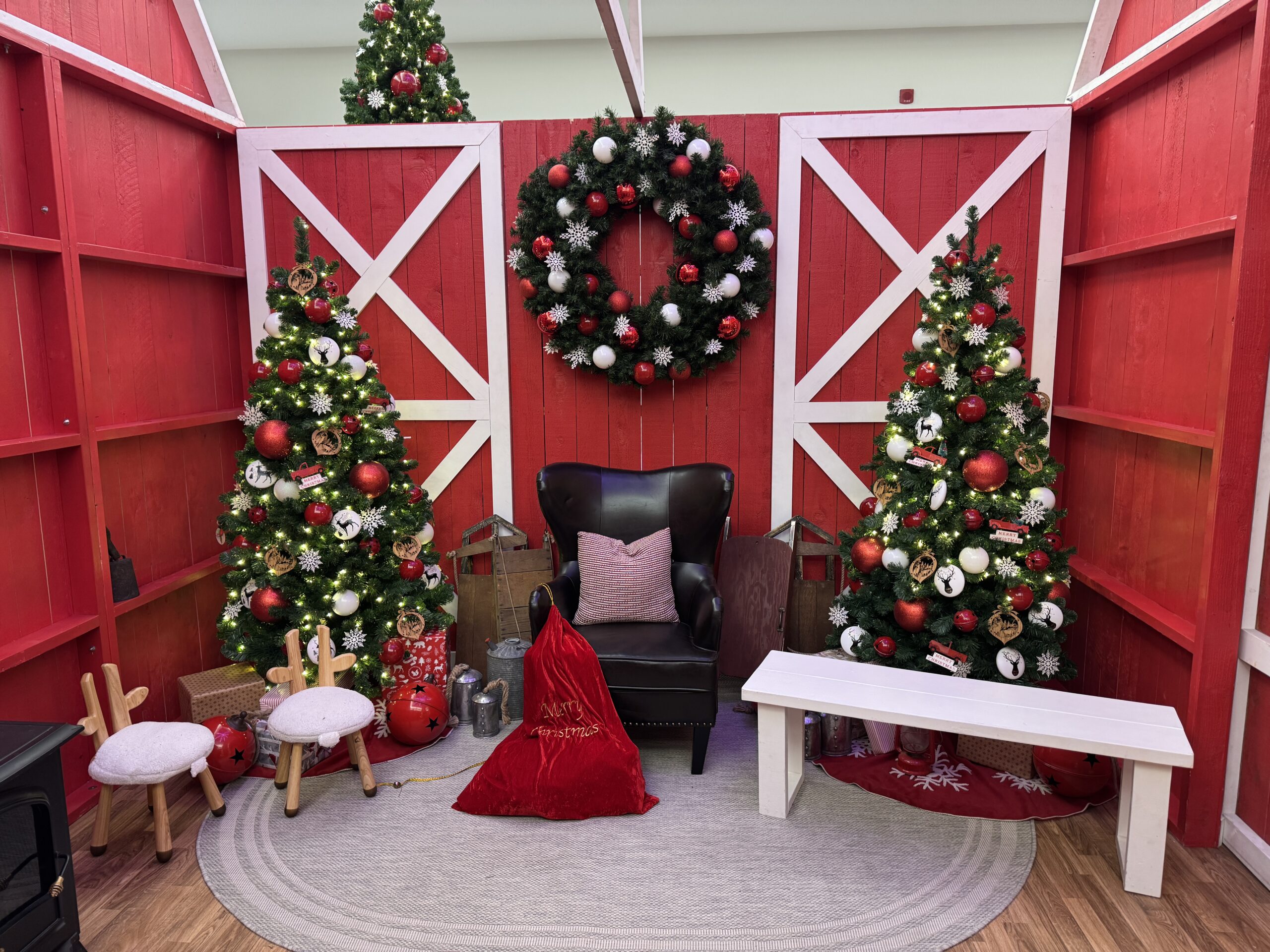 Santa photo set with red barn backdrop, decorated Christmas trees, wreath, and cozy seating in a shopping centre holiday display.