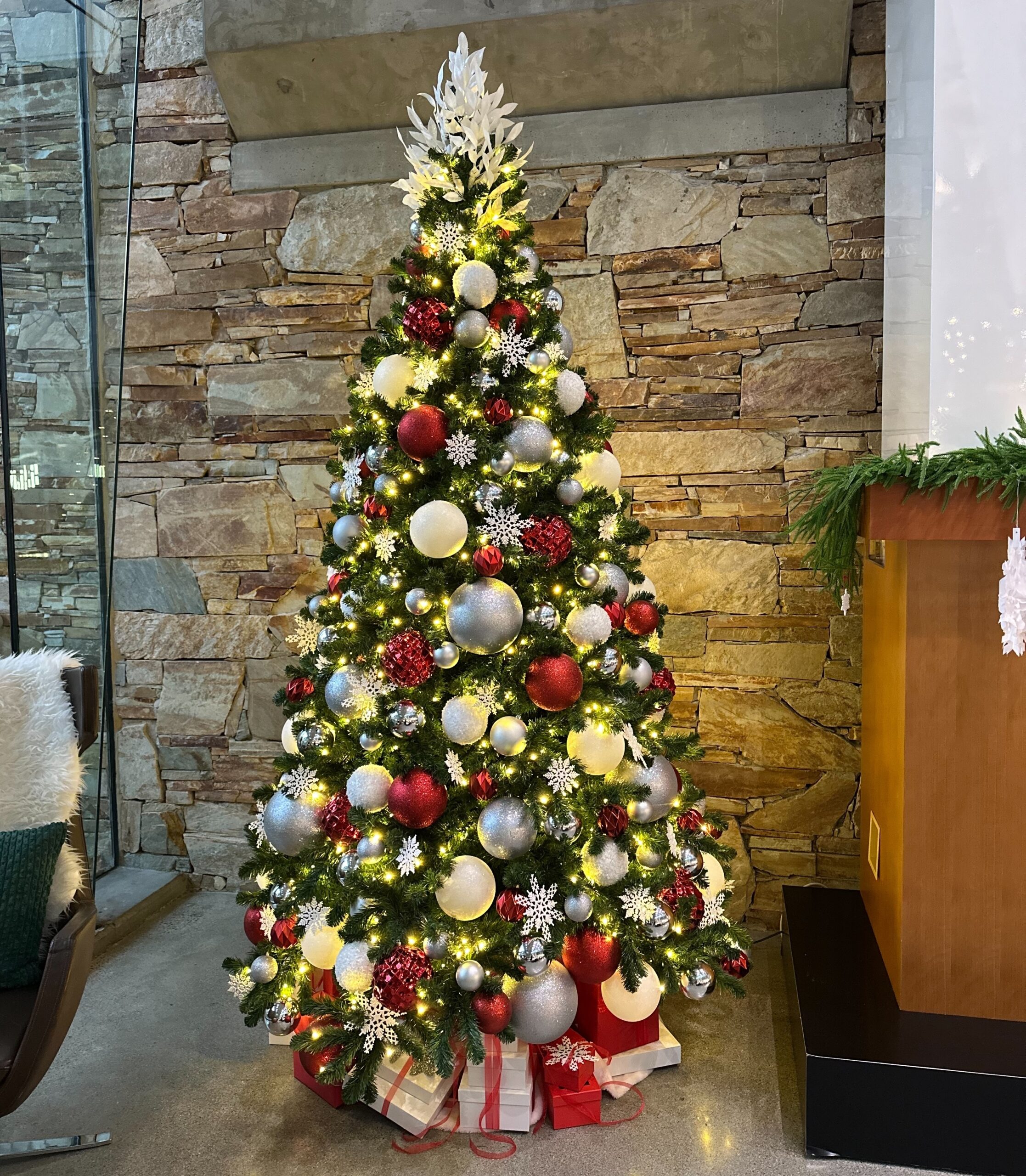 Artificial Christmas tree decorated in red, white, and silver with warm lights, styled in a modern office lobby with a stone feature wall.