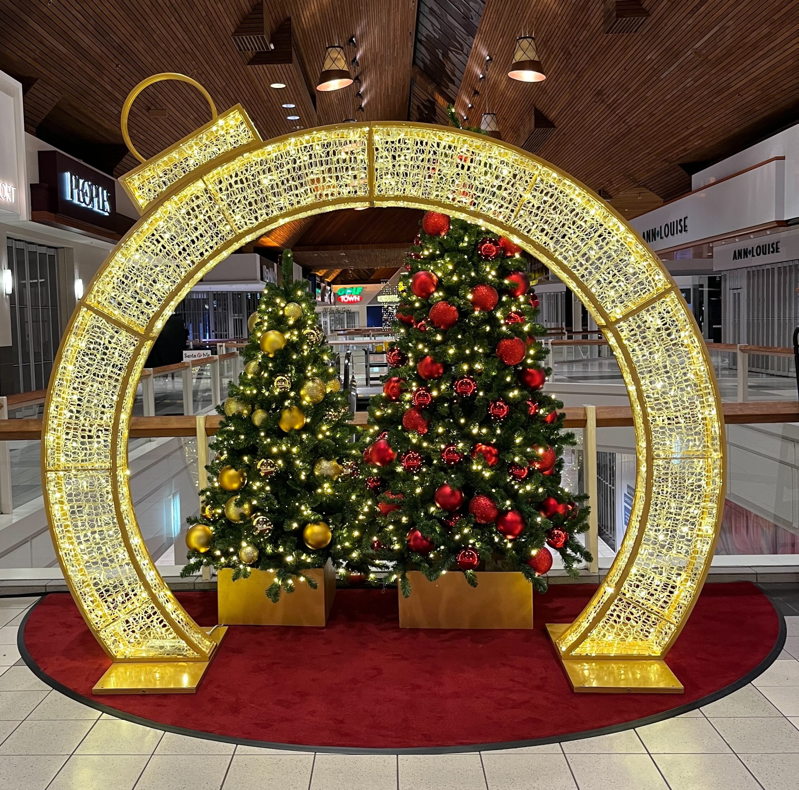 Illuminated gold ornament arch framing red and gold decorated Christmas trees in a shopping centre holiday display.