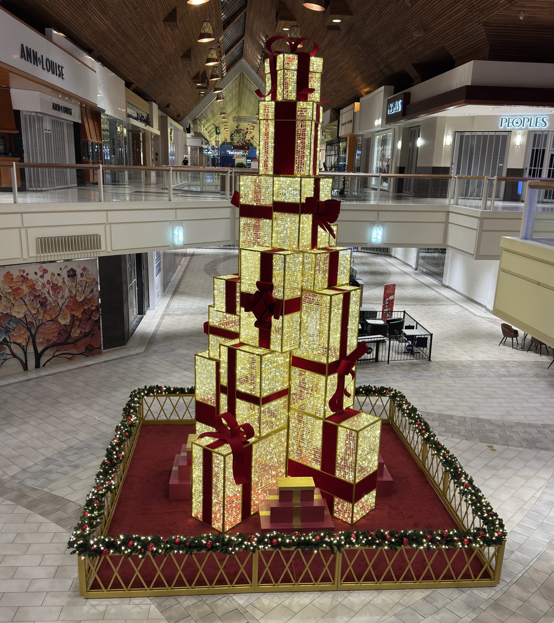 Tall illuminated present stack with glowing gold gift boxes and red ribbon accents in a shopping centre holiday display.