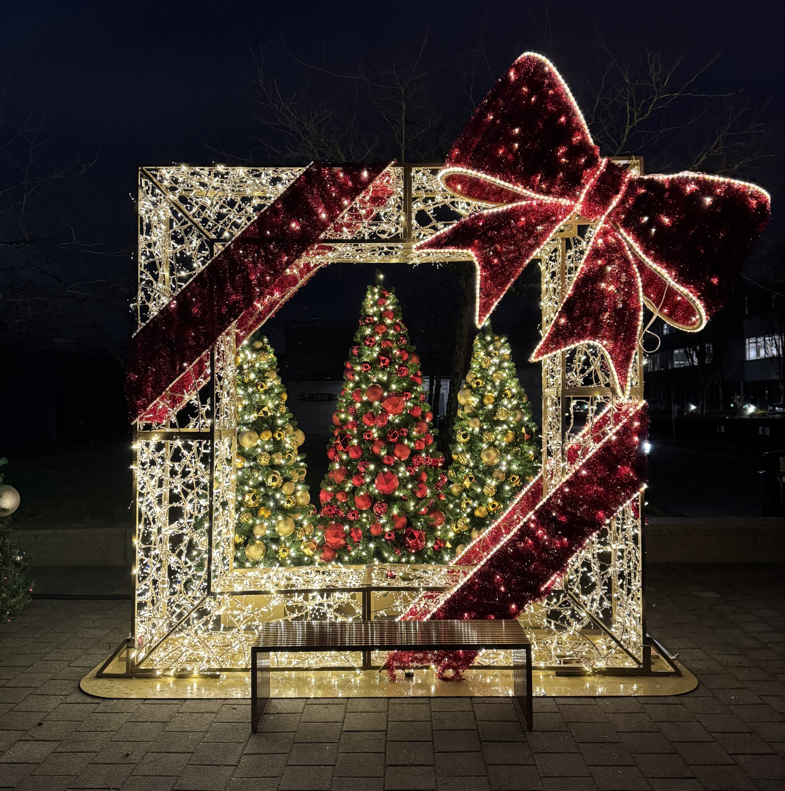 Illuminated outdoor Christmas present structure with red and gold christmas trees in a photo op display at night