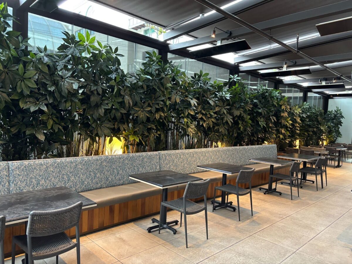 Outdoor restaurant seating with stone bench, small tables, chairs, and tall hedge planters under a covered patio.