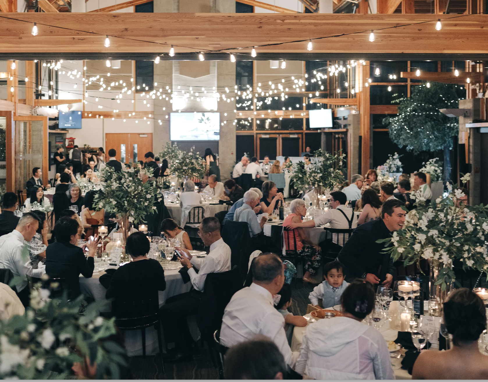 Rustic wedding reception with string lights, artificial green trees and greenery centerpieces