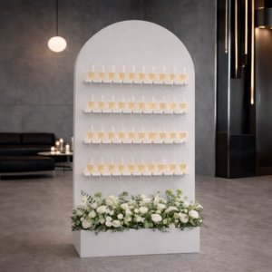 White arched champagne wall display with rows of champagne flutes and floral arrangement at the base for event décor.