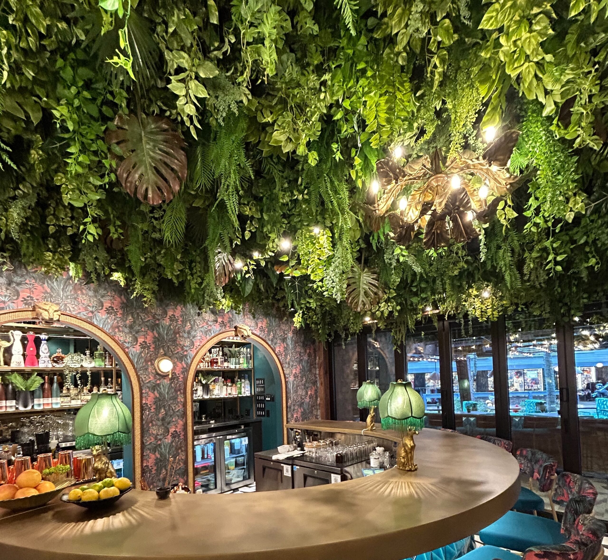 Dense faux hanging greenery canopy with trailing vines, ferns, and large tropical leaves covering a restaurant ceiling above a curved bar.
