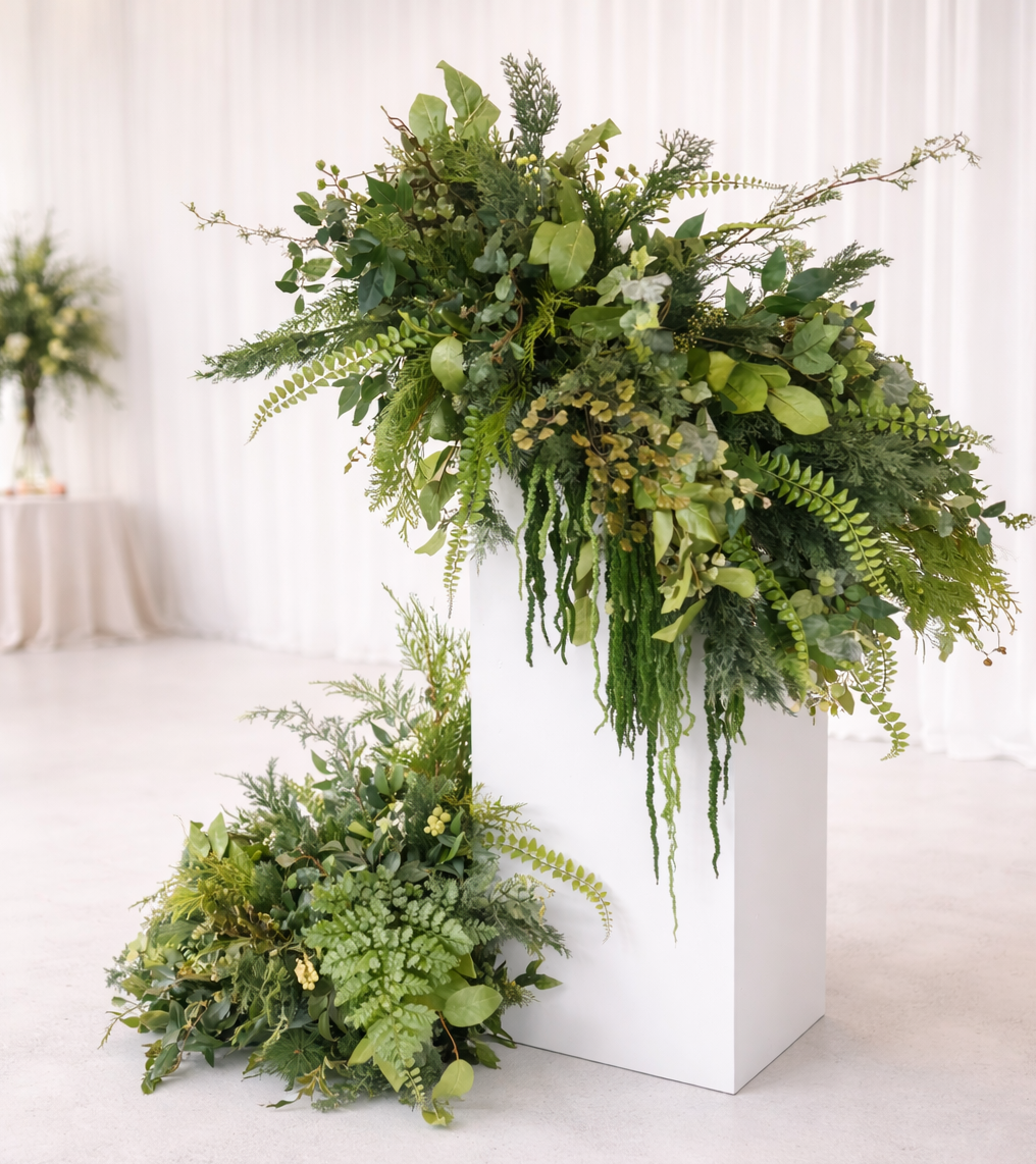 Layered greenery arrangement with mixed faux foliage and trailing stems on a tall white plinth, with a matching floor arrangement in front of white draping.