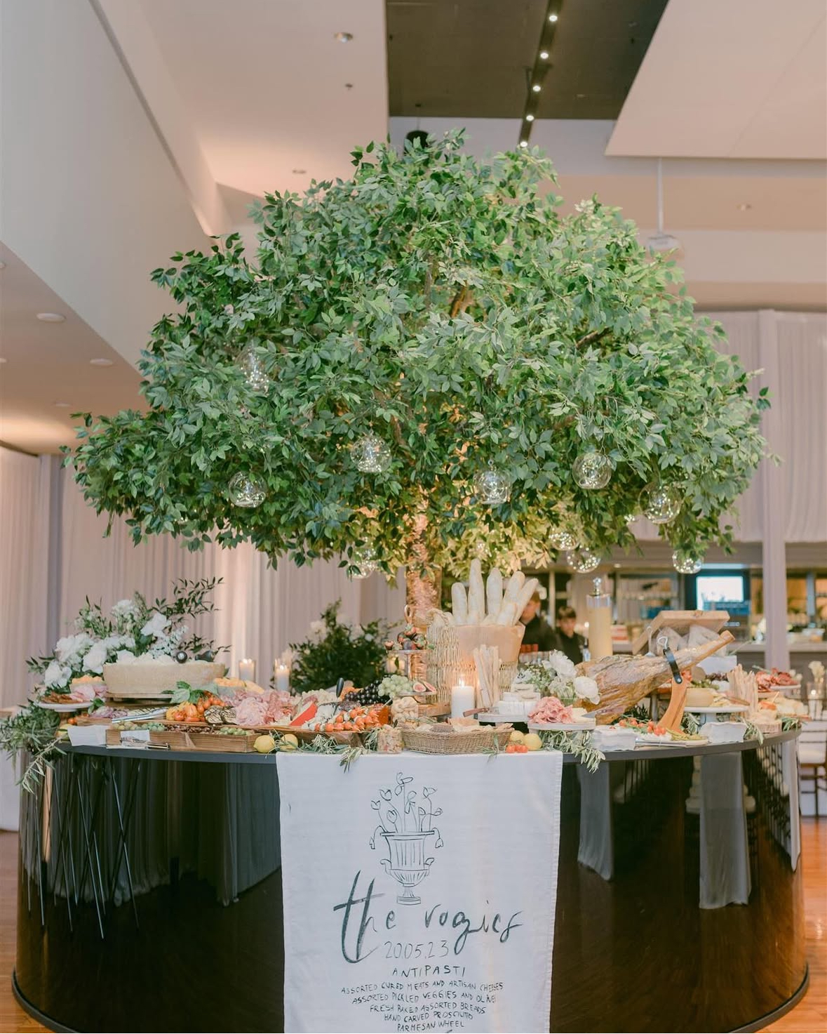 Large canopy tree with dense green foliage and hanging glass orbs above a circular food display table in a corporate event setting.
