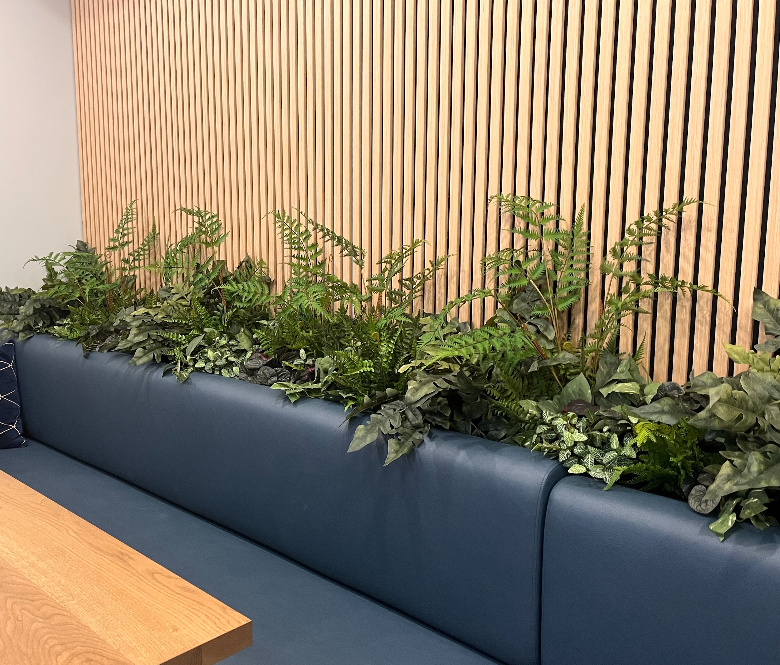 Built-in bench planter filled with artificial ferns and mixed faux greenery along blue office seating against a wood slat wall.