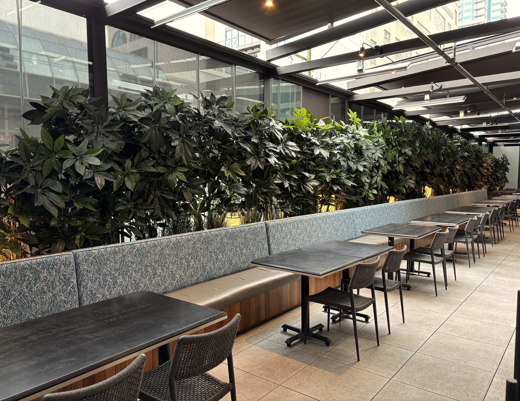 Restaurant patio with banquette seating and tables bordered by a long artificial privacy hedge made of dense faux greenery in a commercial dining space.