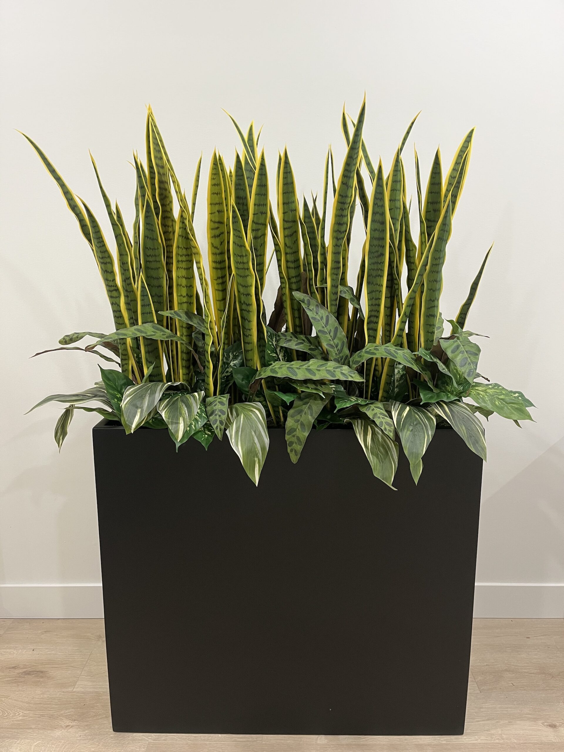 Black rectangular planter with artificial snake plants and mixed faux foliage against a white interior wall.