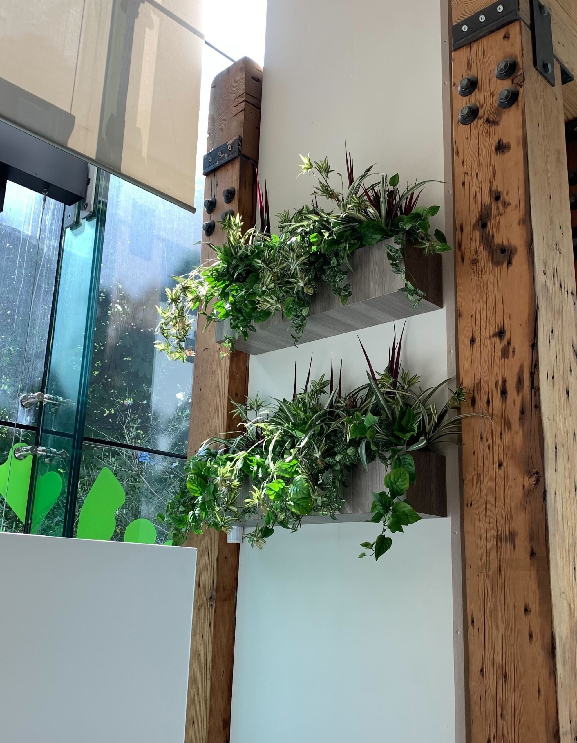 Two rectangular wall-mounted planters filled with mixed artificial greenery and trailing vines on a white wall between exposed wood beams and a glass window