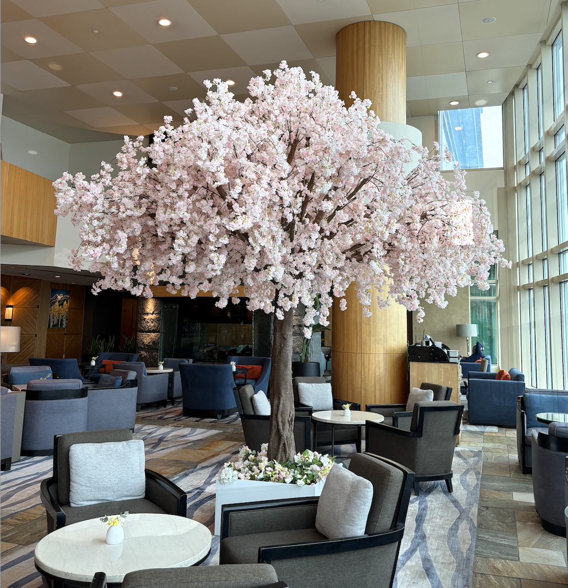 Large artificial cherry blossom tree with pale pink blossoms and a brown trunk in a hotel lobby seating area with lounge chairs and tall windows.