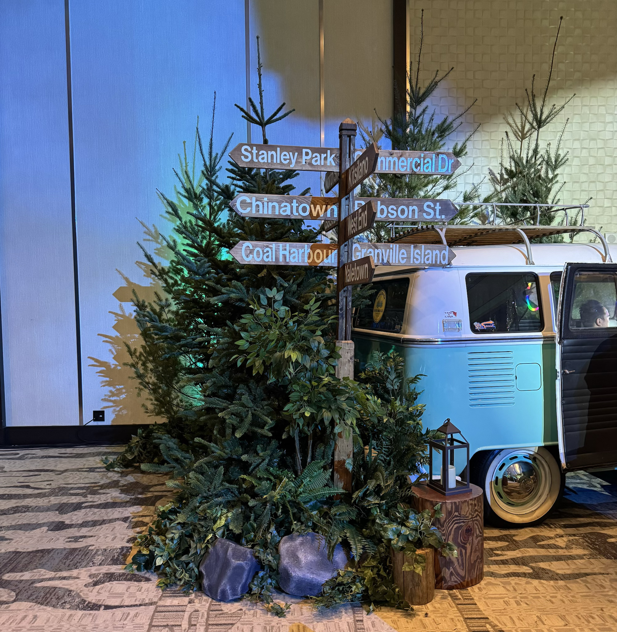 Evergreen trees, mixed greenery, rocks, wood log accents, and a directional signpost beside a vintage van in a corporate event setting.