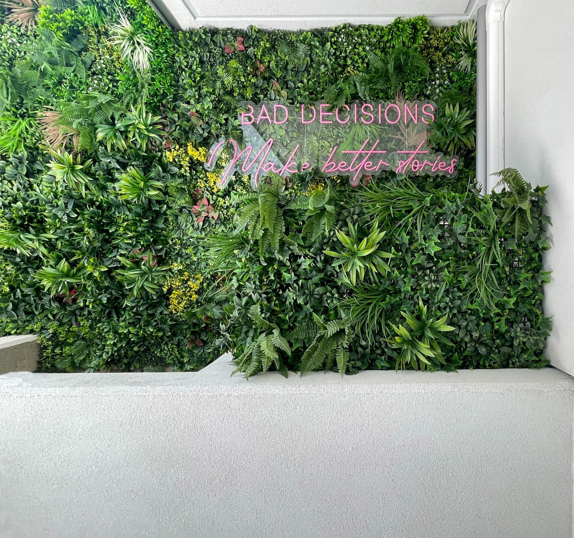 Large faux greenery wall with layered tropical foliage and a pink neon sign mounted in a white commercial interior.