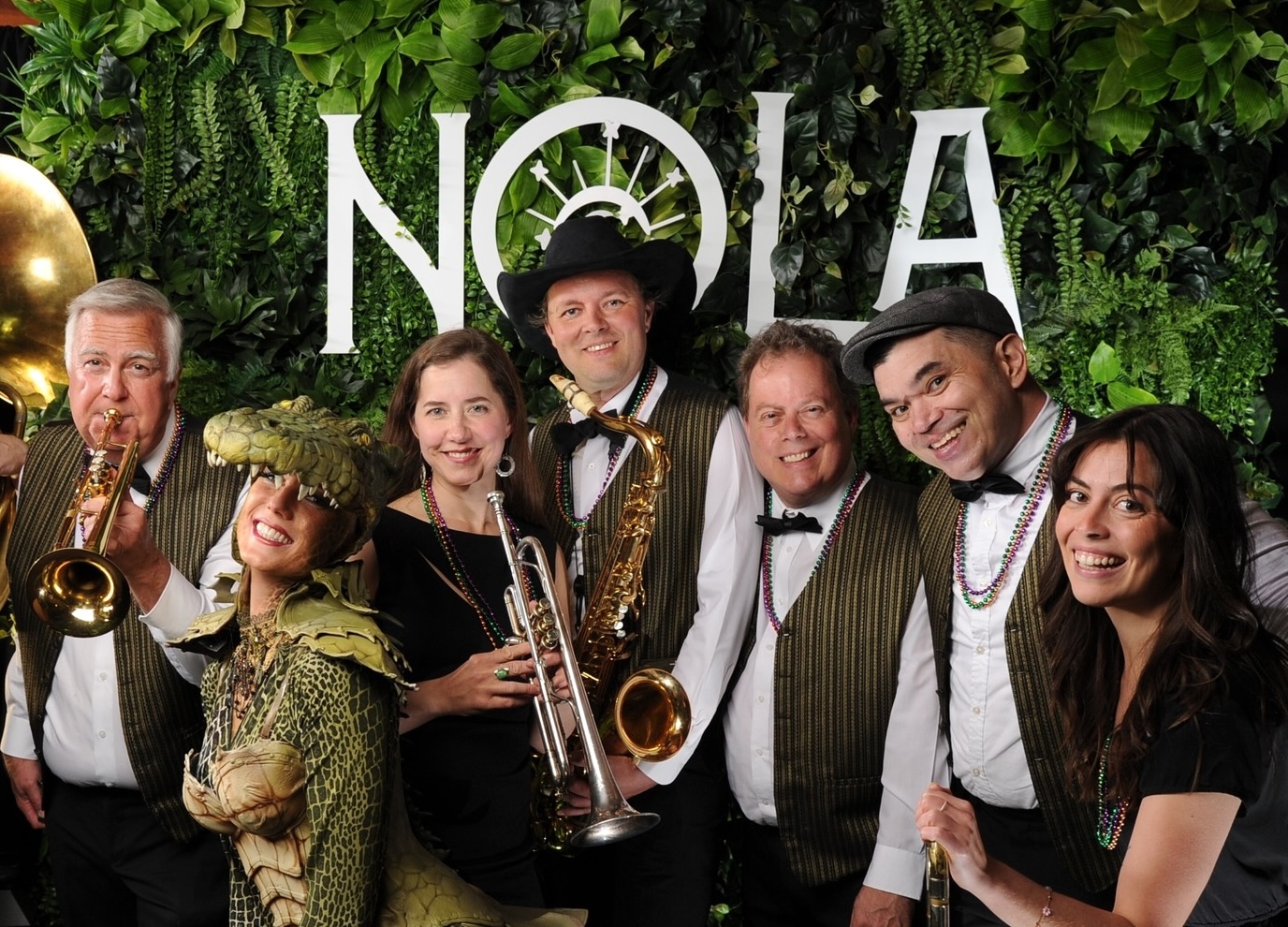 Group posing in front of a textured greenery wall with large white NOLA letters, wearing formal attire and holding brass instruments.